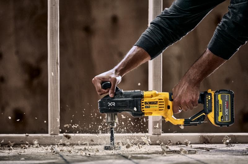 Une personne utilise une perceuse sans fil DEWALT DCD444N avec une poignée latérale pour percer du bois, produisant des copeaux et de la poussière de bois. Une structure de construction est visible en arrière-plan.