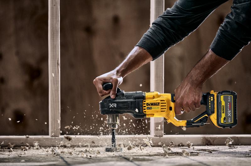 Une personne utilise une perceuse sans fil DEWALT DCD444N avec une poignée latérale pour percer du bois, produisant des copeaux et de la poussière de bois. Une structure de construction est visible en arrière-plan.